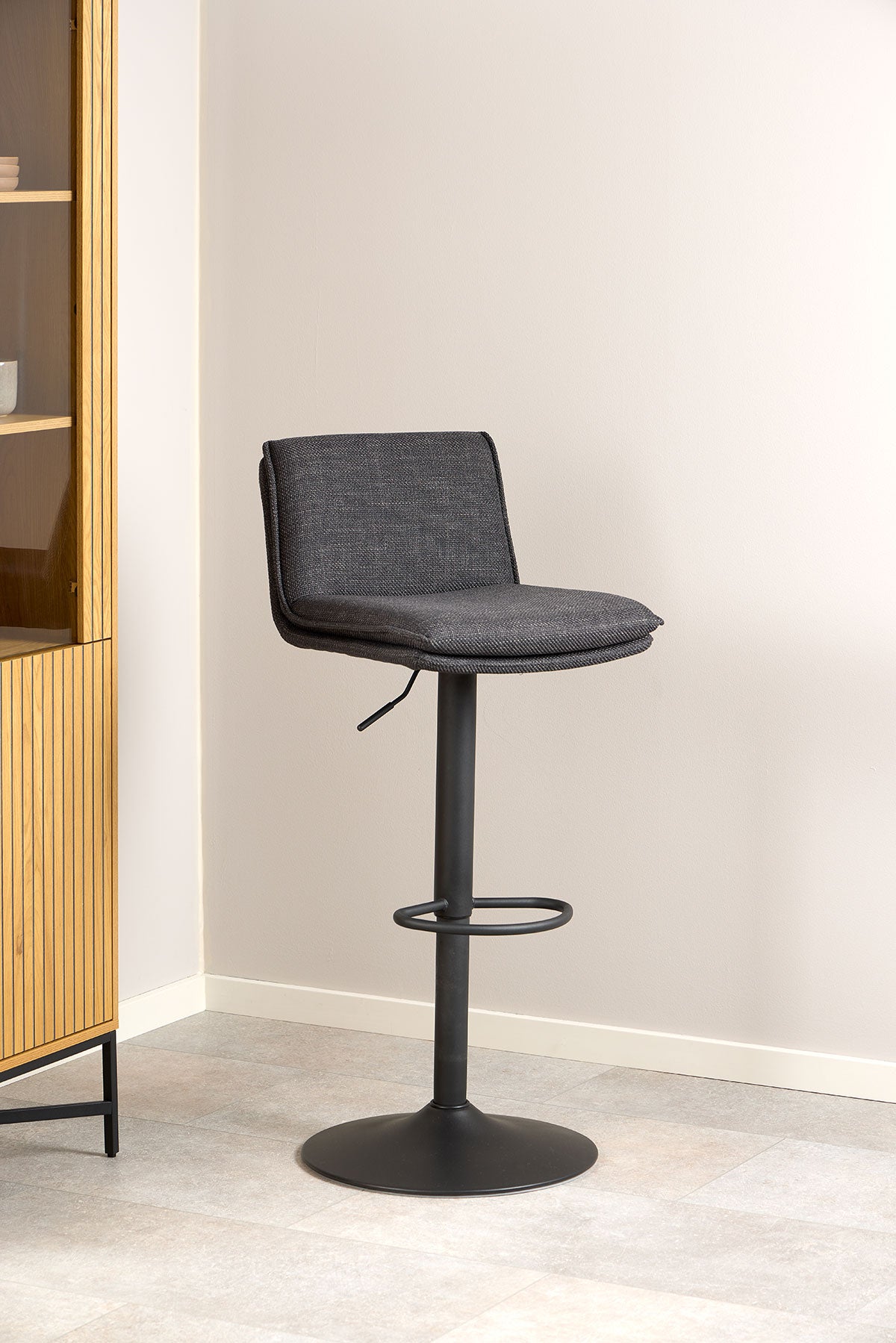 Flynn Barstol i tyget Basel Mörkgrå som står mot en beige vägg
