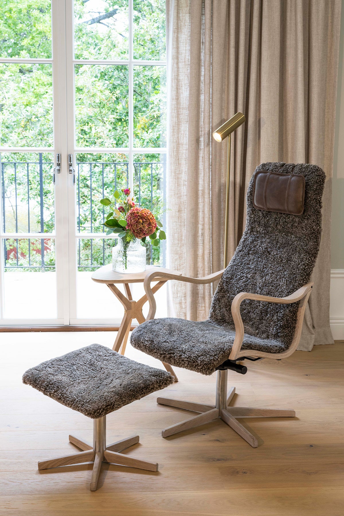 Chic Fåtölj och Fotpall med träfot framför fönster bredvid ett sidobord med blombukett i vas