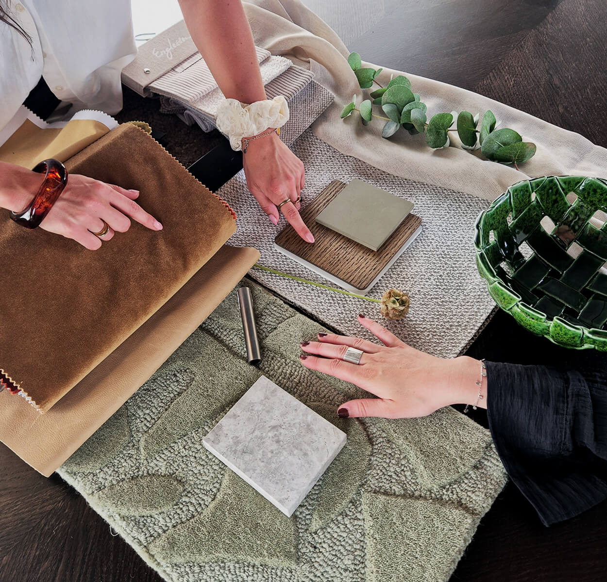 Två personer pekar på olika tyg- och materialprover på ett bord, där en moodboard med textilier, mattor och träslag ligger uppdukad inför val av möbler och inredning.