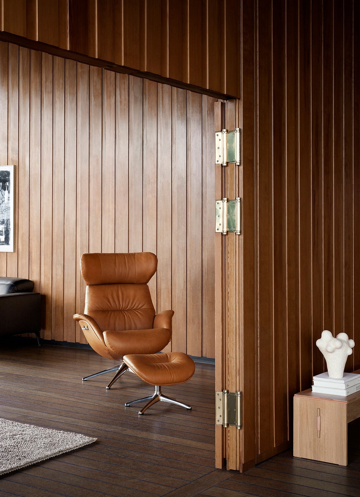 More Fåtölj och fotpall i lädret Dakota Modern Cognac Brown med ben i polerad aluminium i ett rum klätt med träpaneler