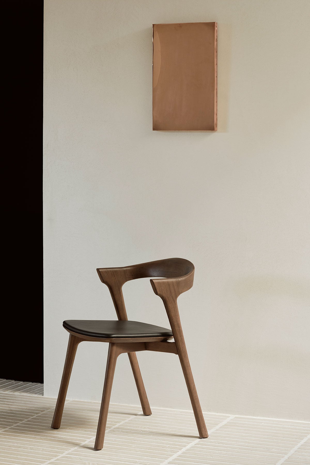 Bok Stol i brunlackad teak med svart lädersits som står mot en vit vägg med en tavla i mässing