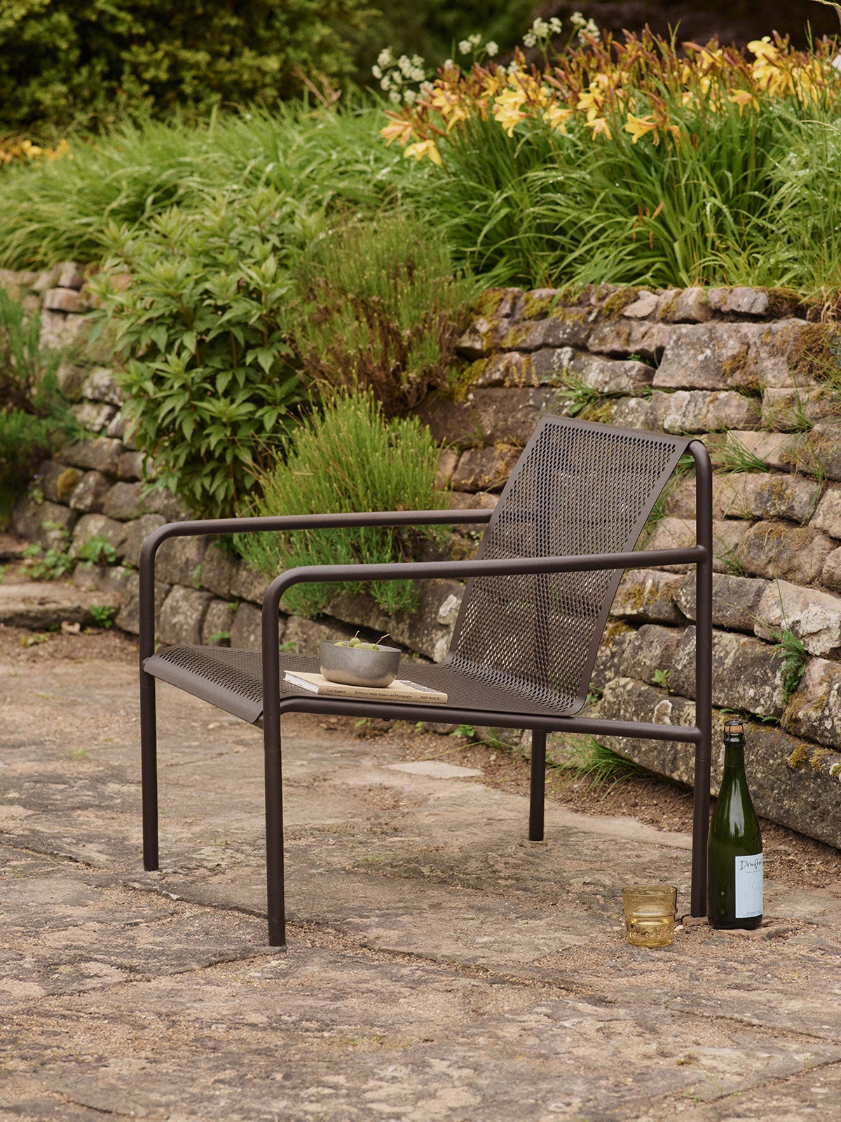 Dapple Loungestol i färgen Dark Chocolate som står på en stenig uteplats fylld med blommor och grönska