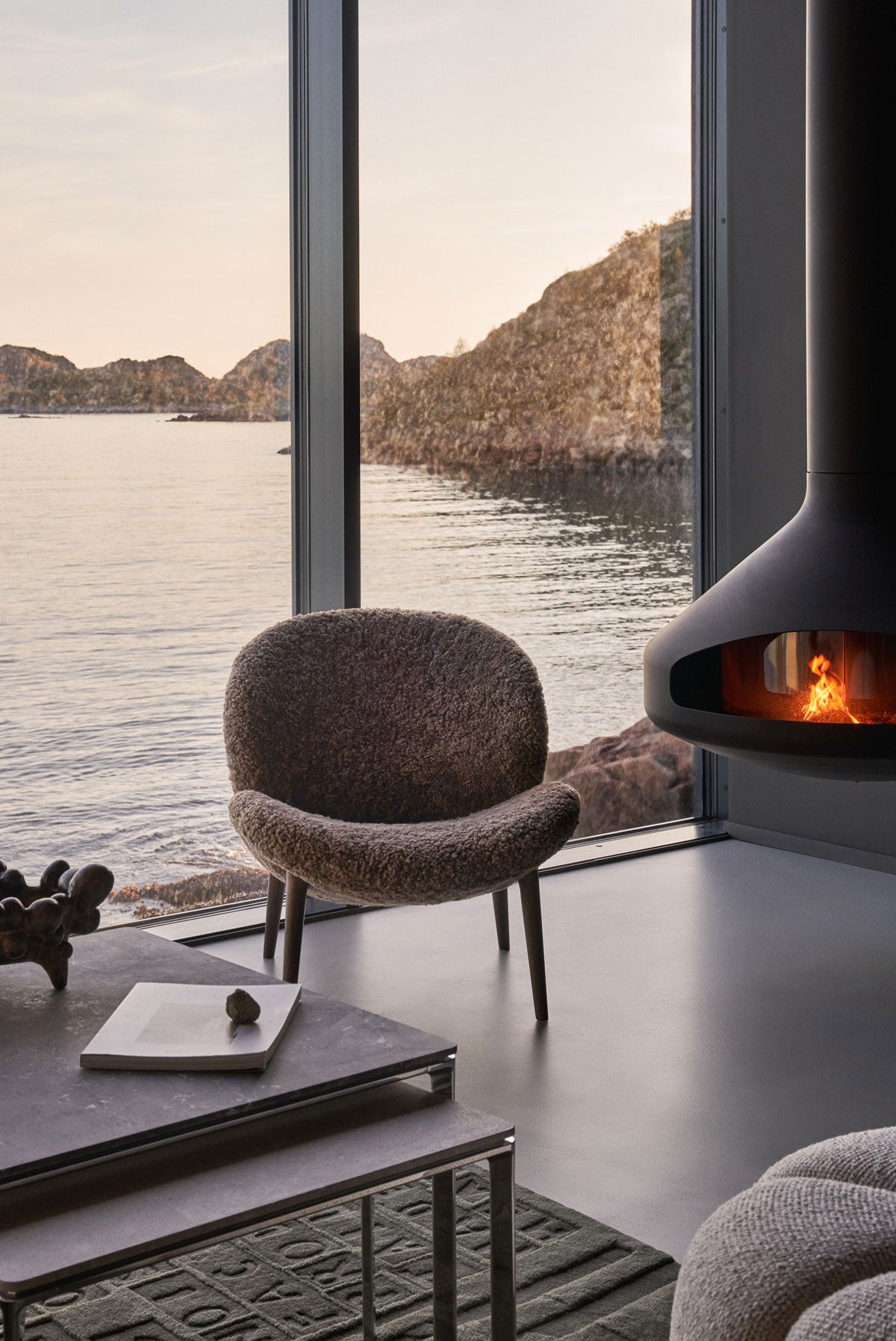 Vipp 469 Lodge Lounge Chair i fårskinn som står i ett vardagsrum framför ett fönster som vetter ut mot en sjö i solnedgången