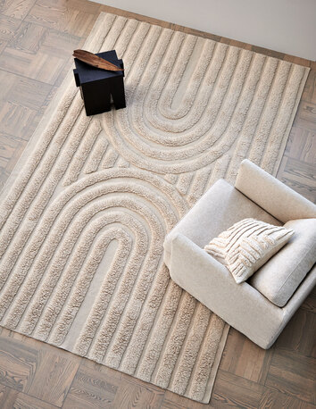 Curve Matta natural beige i ett vardagsrum med mörkt parkettgolv och med en ljus fåtölj på mattan.
