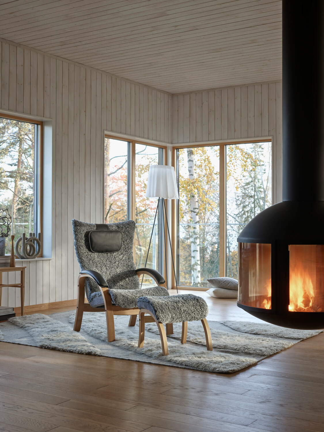 Easy Fåtölj med fotpall i fårskinnet Graphite, bredvid en tänd brasa